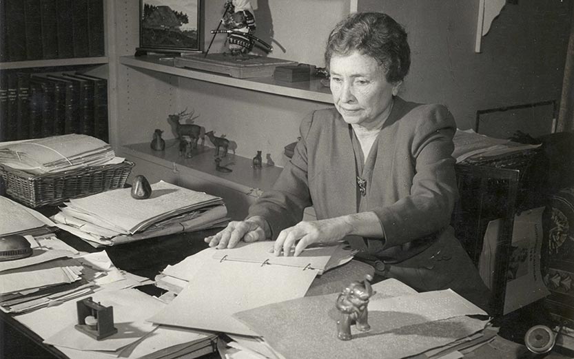 Hellen Keller sitting at a desk while reading braille