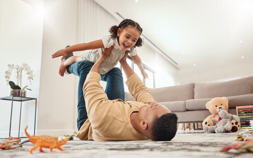 a father laying on the floor lifting his young daughter like an airplane with toys scattered on the floor