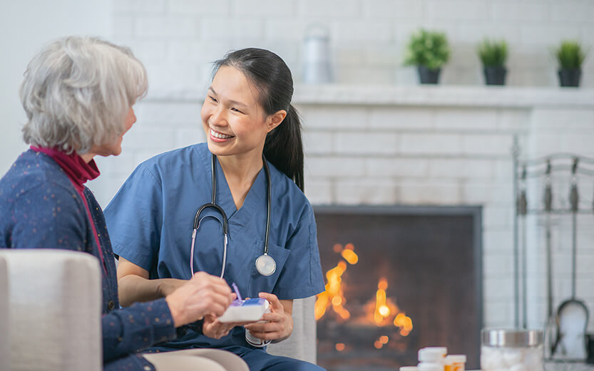 A senior Caucasian woman and an Asian nurse are indoors in a living room. They are talking about the woman's medical regimen.