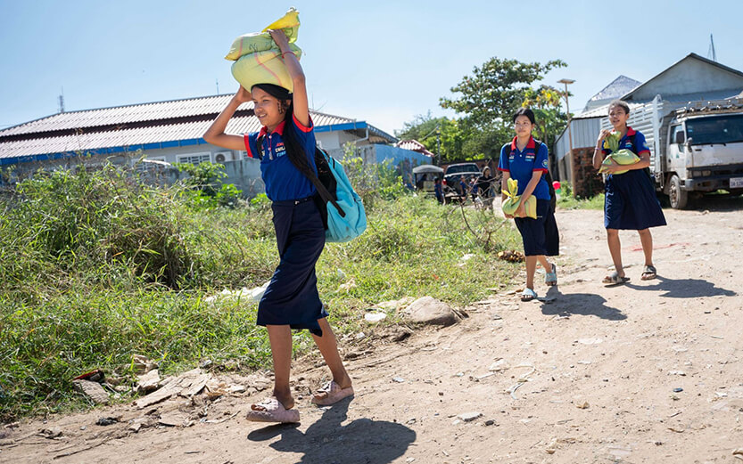 children walking along a dirt road while carrying bags of rice on their heads and in their hands