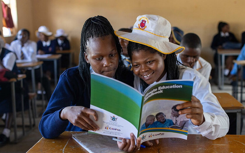 two students sitting at a desk and reading a CAMFED brochure