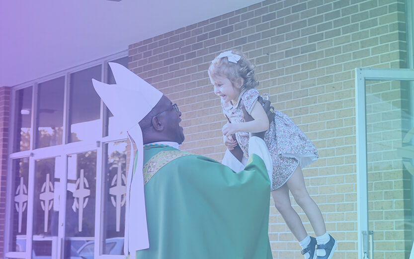 a figure in a green liturgical vestment and white mitre lifting a young child in a floral dress near a brick building