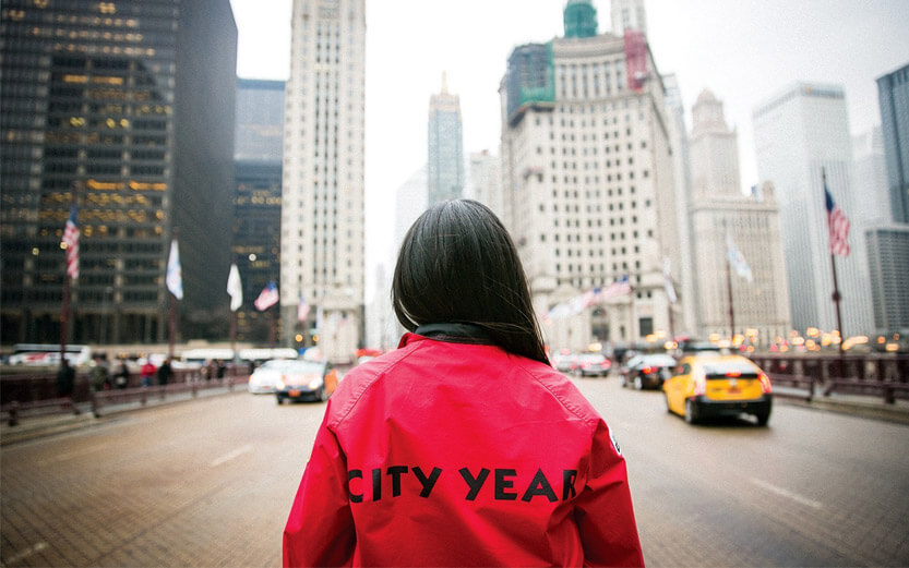 back view of a City Year volunteer wearing a red jacket looking at city buildings