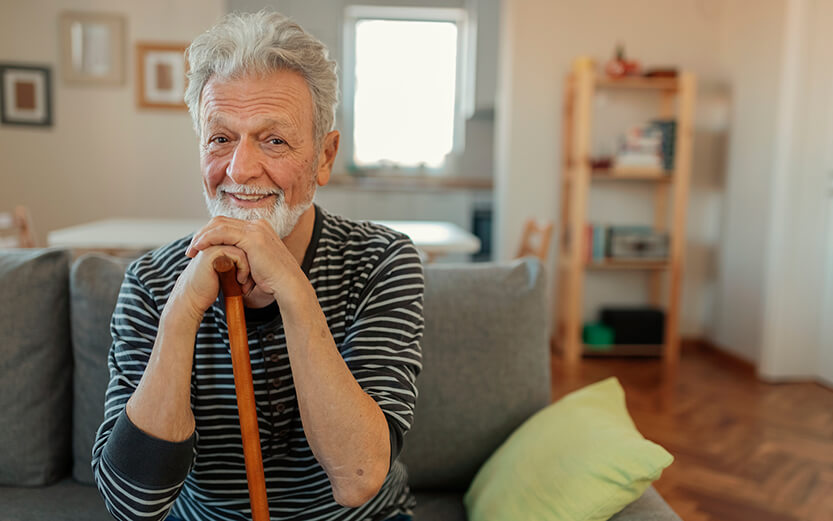 Close up of happy senior man's looking at the camera while holding the walking cane, he is sitting on the sofa in the living room