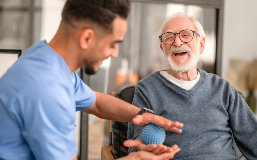 a senior man laughing while his physical therapist works on his hands with a nubbed ball