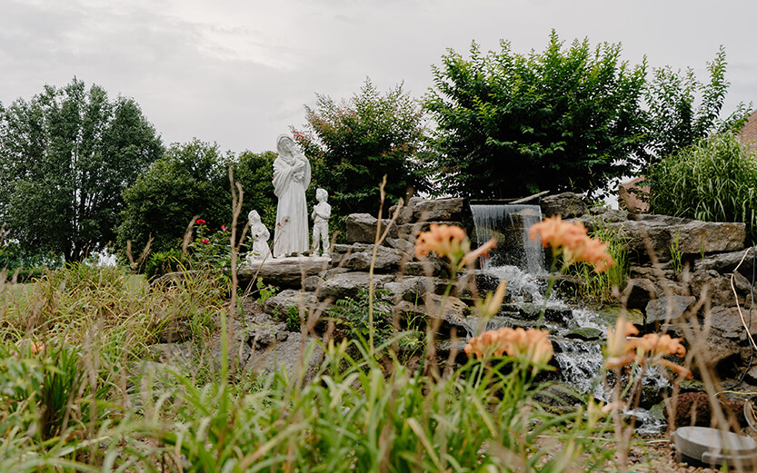 a religious outdoor grotto and waterfall garden featuring white statues of the Virgin Mary holding the Child Jesus, accompanied by kneeling and standing figures next to a stone rock waterfall and orange daylilies