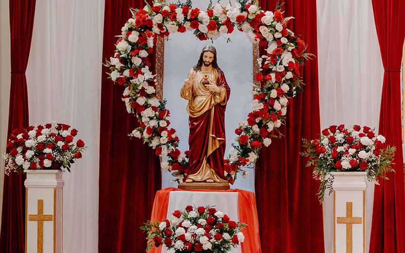 a statue of the Sacred Heart of Jesus set up on a church altar decorated with a large heart-shaped arrangement of red and white roses, white and red drapery, and floral pedestals