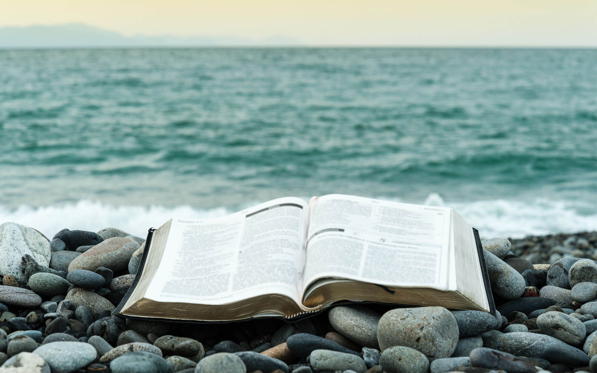 Bible Open On Top Of Small Stones In Front Of The Green Sea