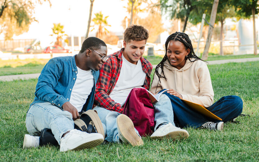 three students sitting on a lawn looking at a book