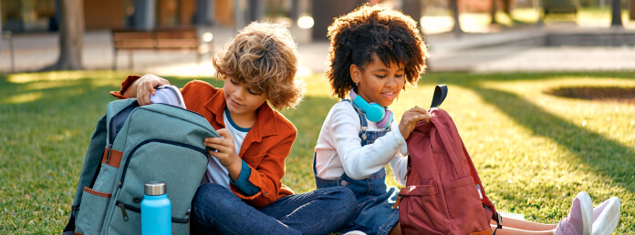 2 children sitting on the grass looking into their backpacks