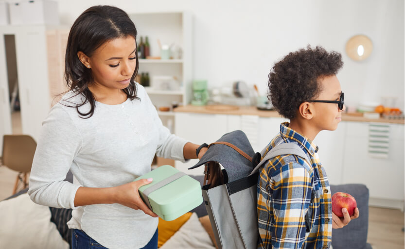 a mother packing her son's lunch bag into his backpack