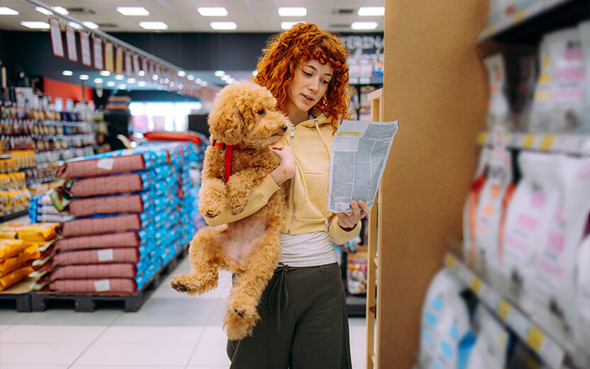 A person with curly red hair holds a small brown dog while reading a product label in a pet store aisle