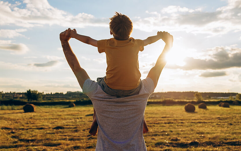 Father carrying son on shoulders in a sunny countryside field, holding hands and enjoying a joyful summer moment