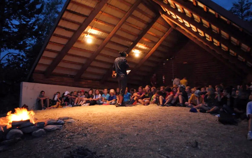 a camp leader talking to a group of campers in the evening under an A-frame shelter