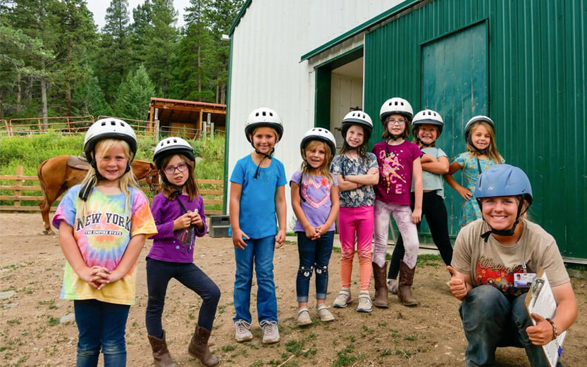 a group of young girl campers wearing white helmets getting ready to ride horses