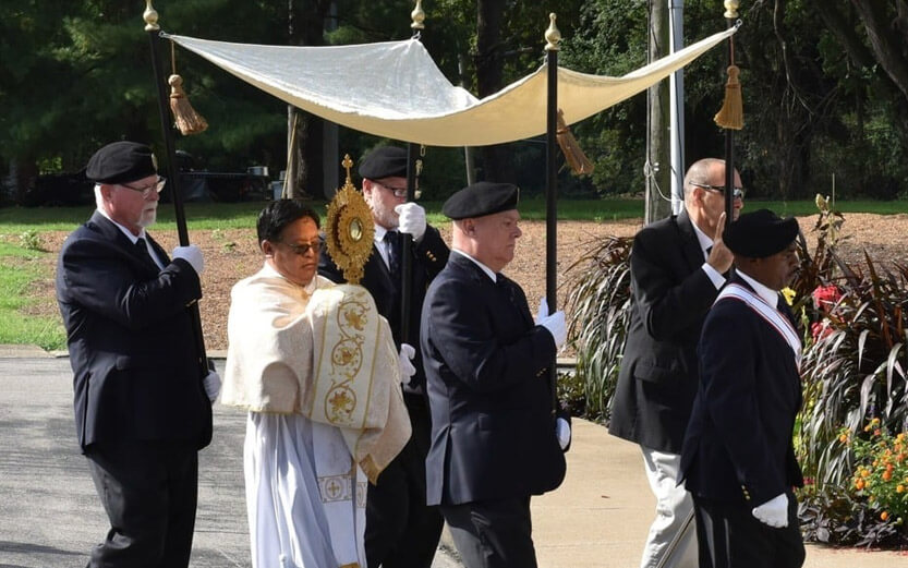 a procession with a pastor carrying a cross while men carry a canopy to cover them