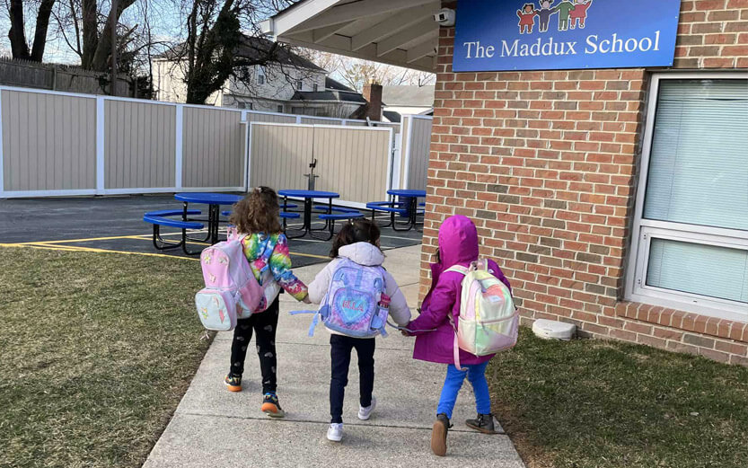view from behind of three preschoolers holding hands walking to their school building