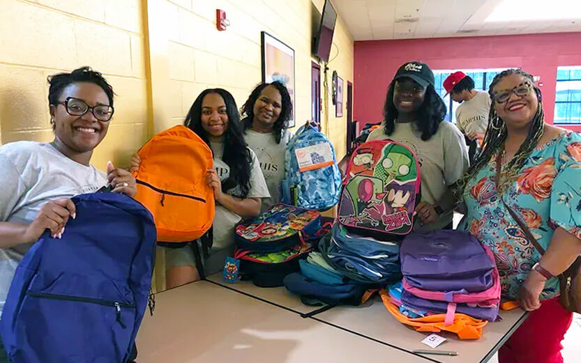 five volunteers with a stack of backpacks to be donated