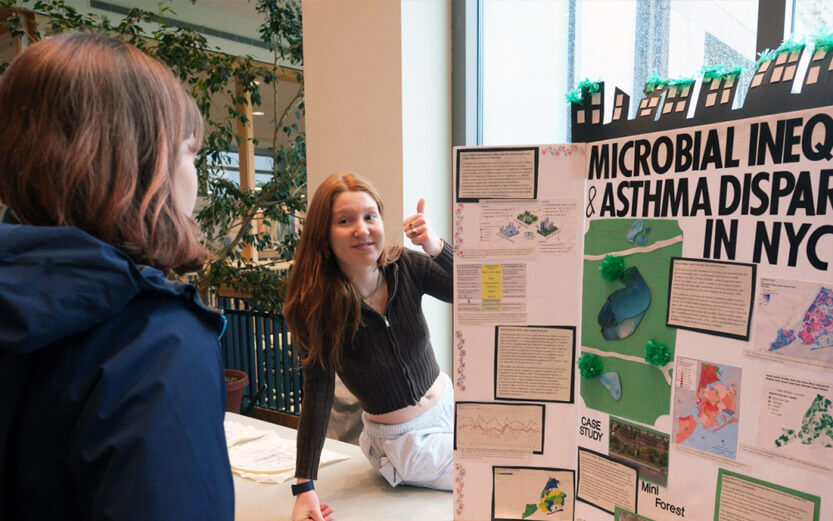 a student sitting next to her science presentation board and giving the thumbs up sign