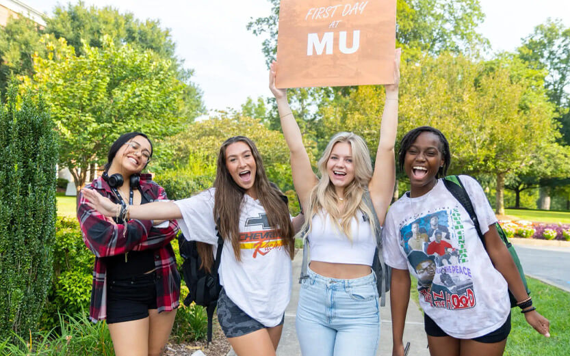 four female students smiling and holding a sign that reads First Day at MU