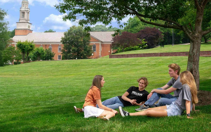four students sitting on the school lawn under a tree with a school building in the background