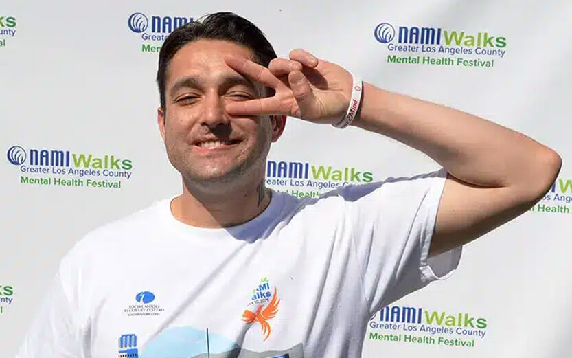 a man in front of a NAMIWalks step and repeat display smiling and flashing a peace sign