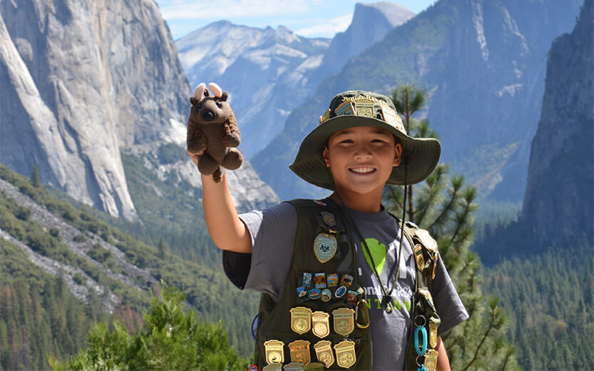 A young Junior Ranger smiles broadly while posing in front of a scenic mountain valley view at Yosemite National Park. He wears an outdoor sun hat and a vest completely covered in official ranger badges and pins while holding up a small plush bison toy.