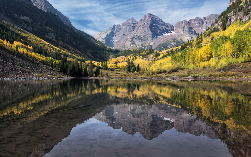 A scenic mountain landscape featuring the Maroon Bells peaks perfectly reflected in the calm waters of Maroon Lake. The slopes surrounding the lake are covered in vibrant yellow autumn aspen trees and green evergreens under a partly cloudy blue sky.