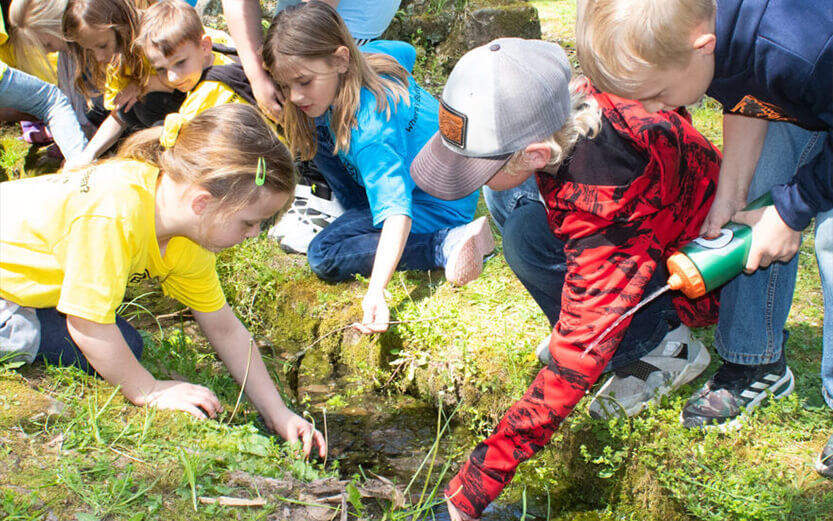 A group of young children gathers outdoors around a small stream on a grassy bank. A girl in a yellow shirt and a girl in a blue shirt lean forward to touch the water, while a boy in a red and black hoodie squirts a stream of water into the creek from a green squeeze bottle.