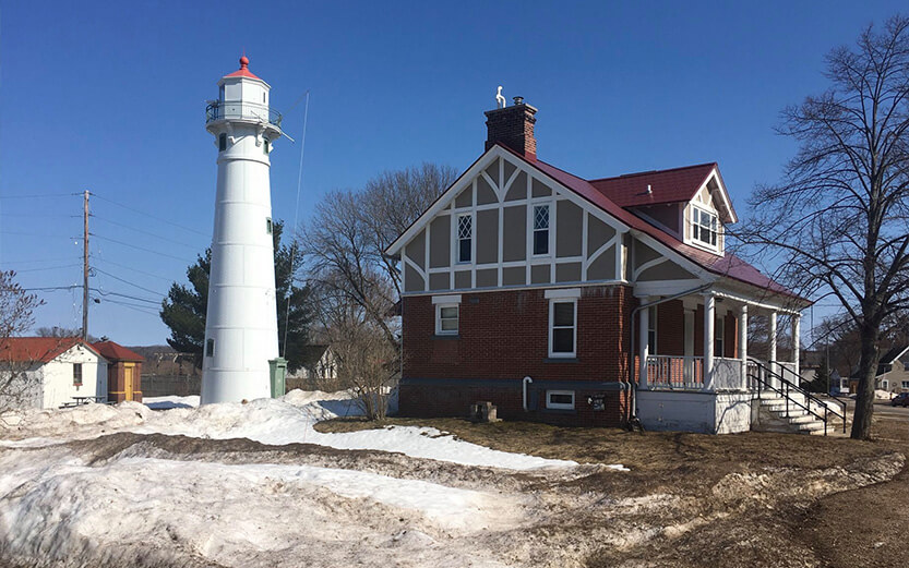 A white, cylindrical lighthouse with a red roof stands next to a two-story brick keeper's house featuring a gabled roof and half-timbered styling. Patches of melting, dirty snow cover the ground under a clear blue sky.