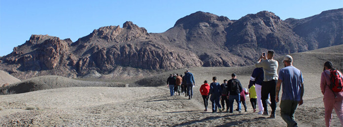 A large group of people hikes in a single-file line along a dirt trail winding through a dry, barren desert landscape. Towering, rugged brown rock mountains rise up in the background under a clear, bright blue sky.