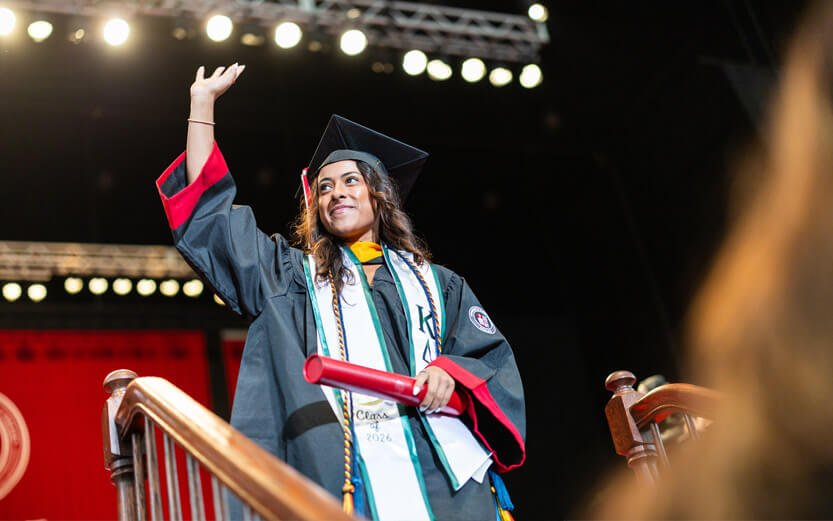 a graduate on stage holding her diploma smiling and waving