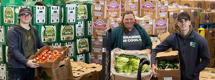 three volunteers showing off boxes of produce in a food bank warehouse