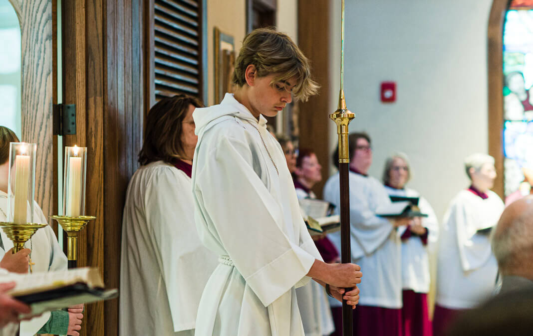 teen boy wearing a white robe, holding a large staff with a cross at the back of the church
