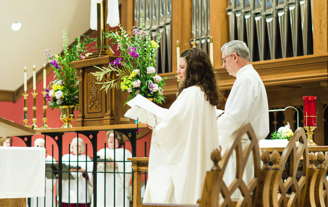 woman and man in white robes at the front of the church reading scripture