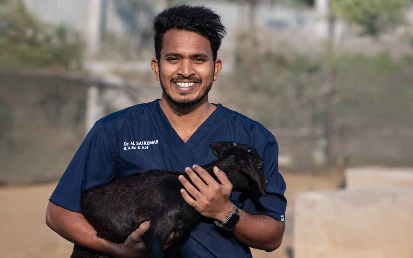 a smiling veterinarian in blue uniform holding a black baby goat