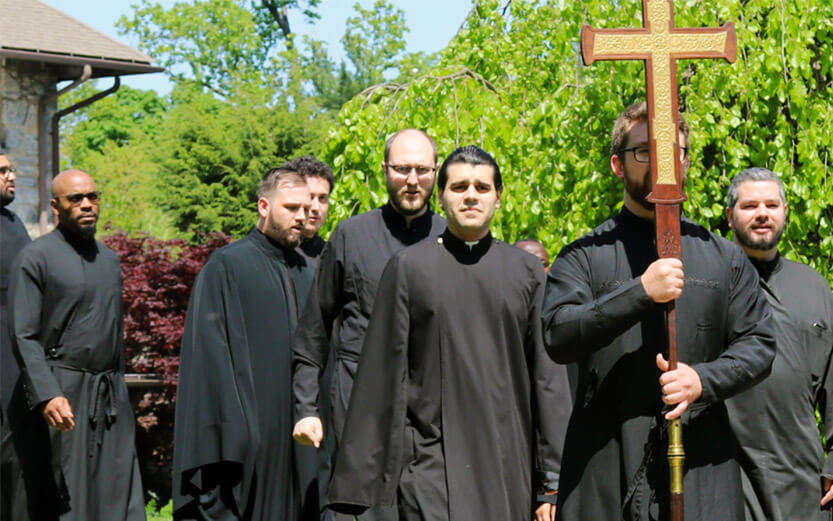 a procession of church clergy in black robes with the person in front carrying a cross