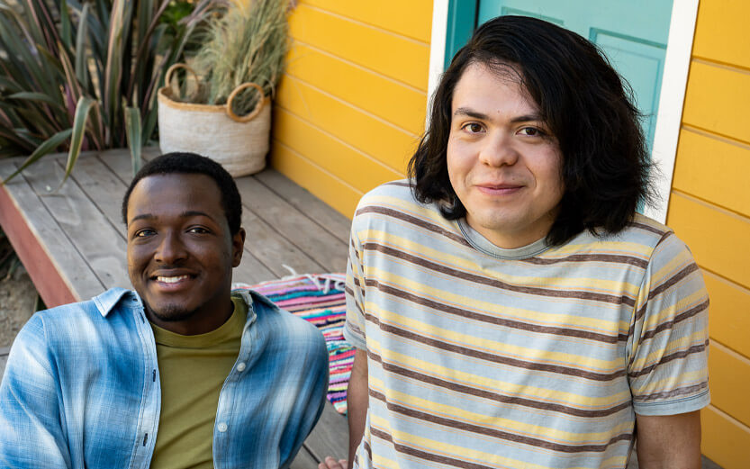 two LGBTQ+ people sitting in front of a house smiling