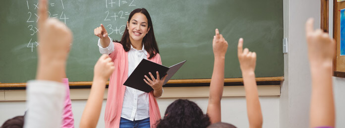 a teacher standing in front of a classroom with students hands raised in foreground
