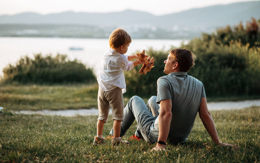 a father sitting on the grass while his son plays with a wooden airplane
