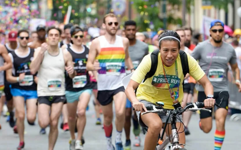 a woman riding a bike in front of the pack of runners at a Pride Run